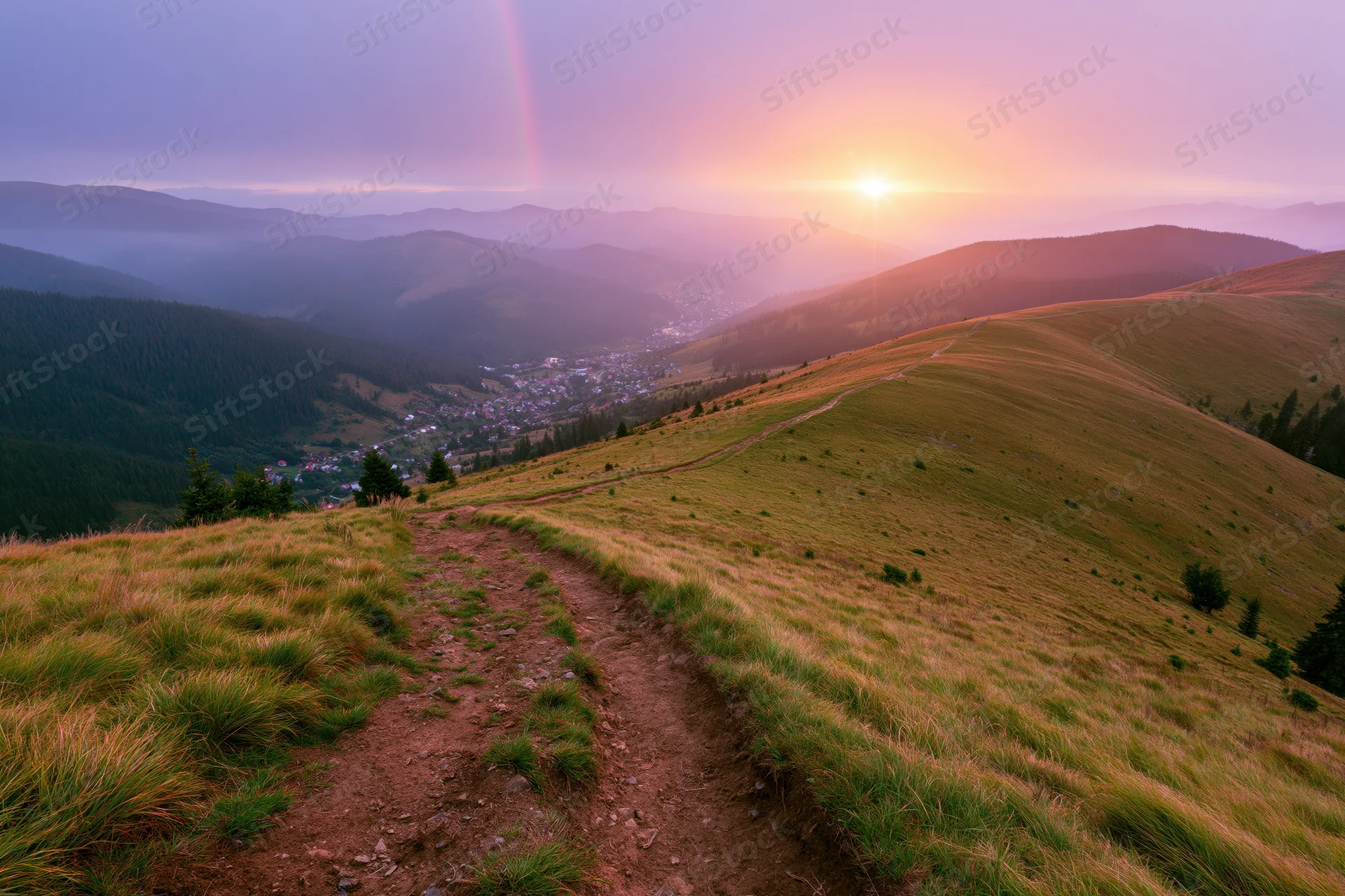 Dirt path curving down a grassy mountain hillside toward a valley village at sunset