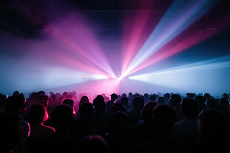 Dense crowd of silhouetted people facing a stage with colorful laser beams in an indoor concert venue