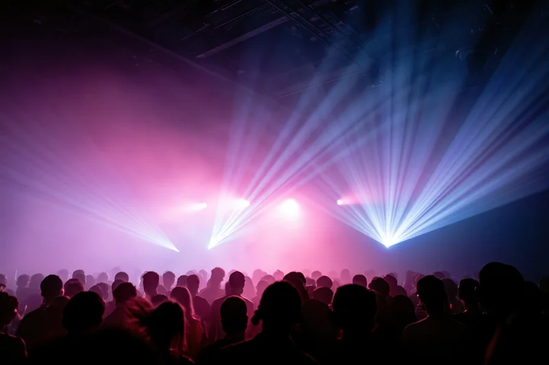 Dense crowd of silhouetted people facing a stage with colorful laser lights and smoke at an indoor concert event