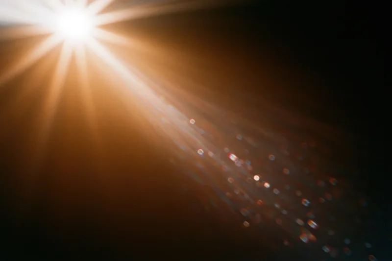 Intense lens flare from a bright light source casting radiant beams and sparkling particles across a dark background