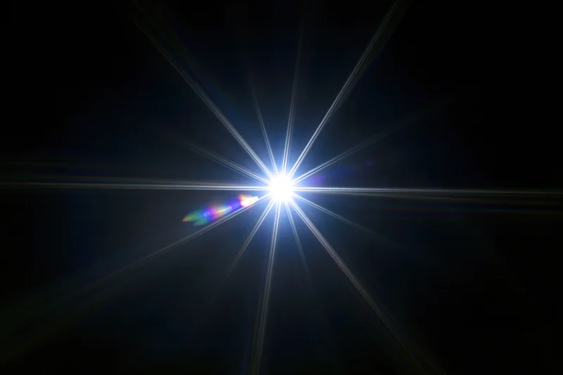 Intense white starburst lens flare centered against a pure black background with radial light beams and chromatic dispersion on one side
