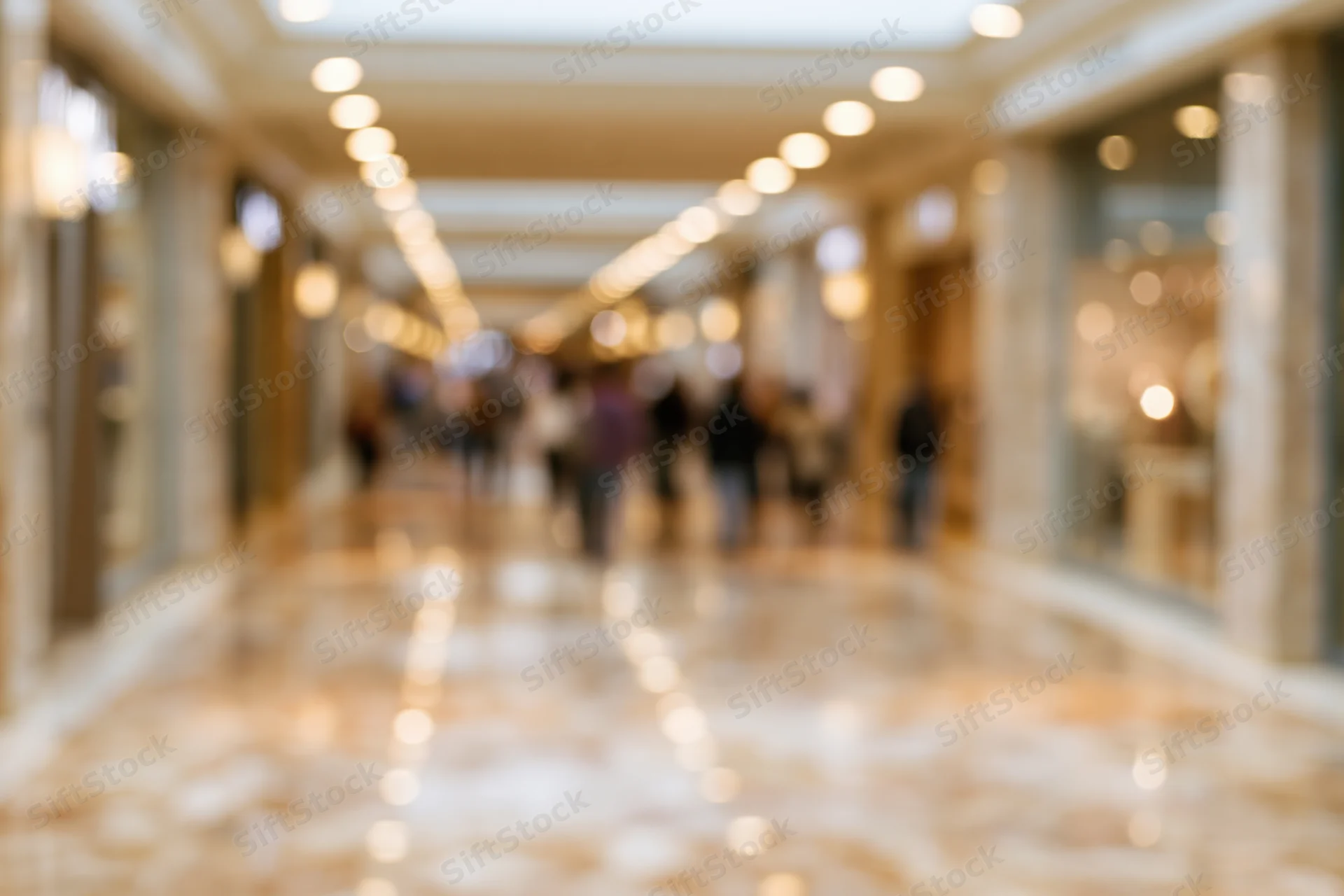 Defocused interior of an upscale shopping mall with blurred people walking along a marble-floored corridor lined with storefronts and recessed ceiling lights