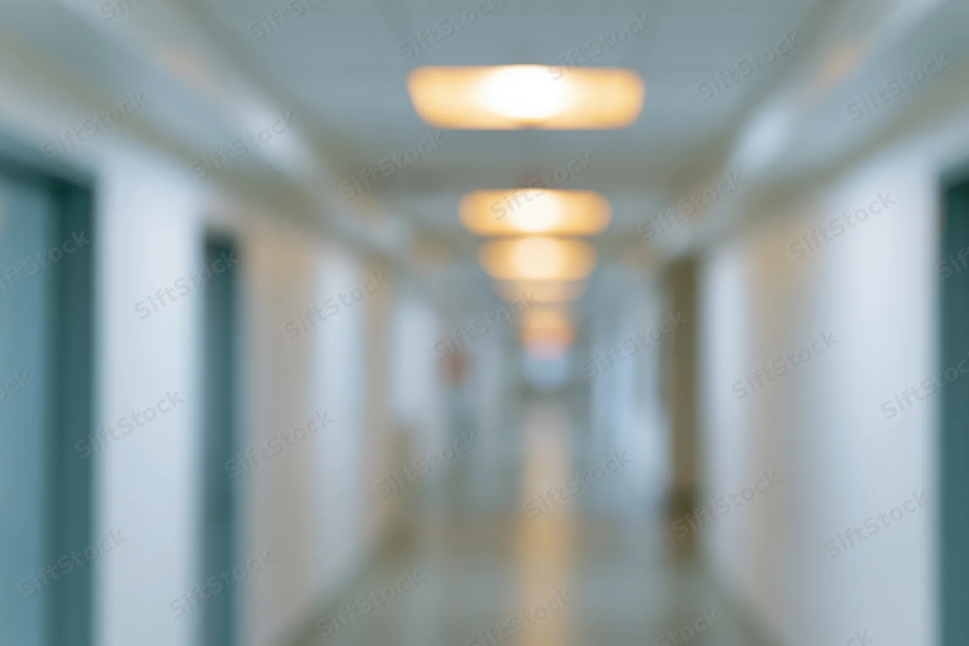 Defocused view down a long empty hospital corridor with ceiling lights and doors on either side