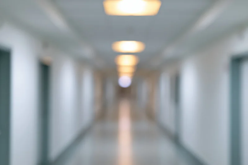 Defocused view down a long empty hospital hallway with glowing ceiling lights and doors along the sides