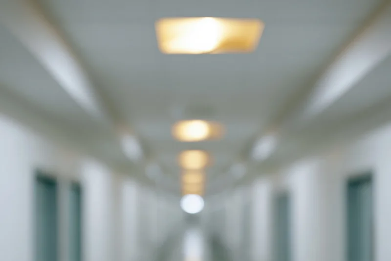 Defocused view down a long empty hospital hallway with ceiling lights and white walls