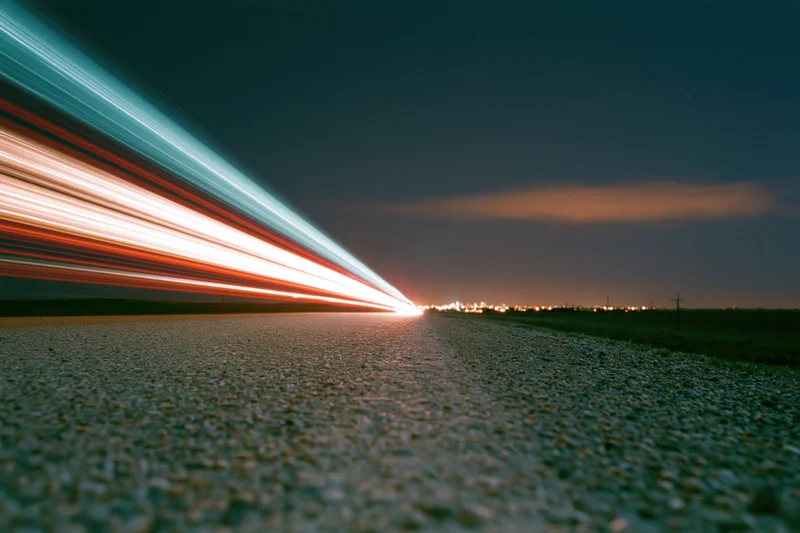 Long-exposure light trails from vehicles moving along a dark asphalt road at night toward a distant city skyline
