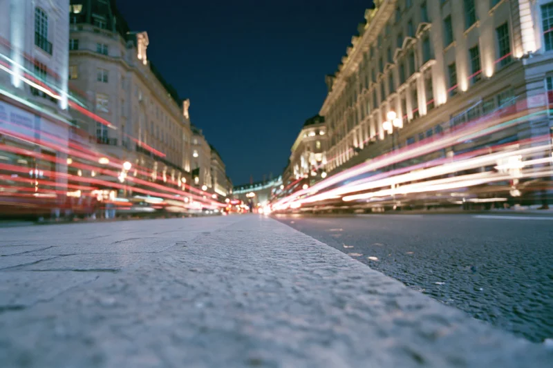 Long-exposure night view of a busy city street with light trails from moving vehicles along a cobblestone sidewalk and illuminated classical buildings
