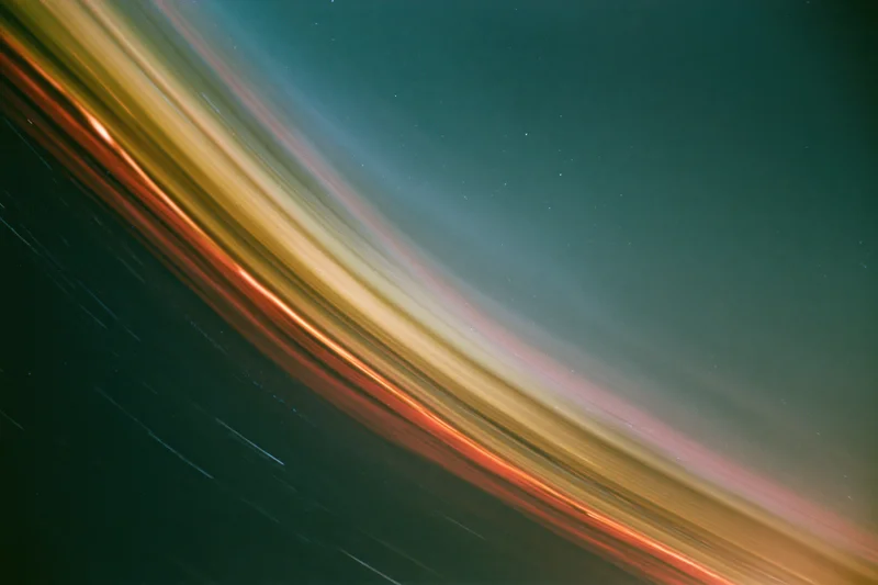 Long-exposure night sky with diagonal star streaks across dark teal and warm-toned gradients