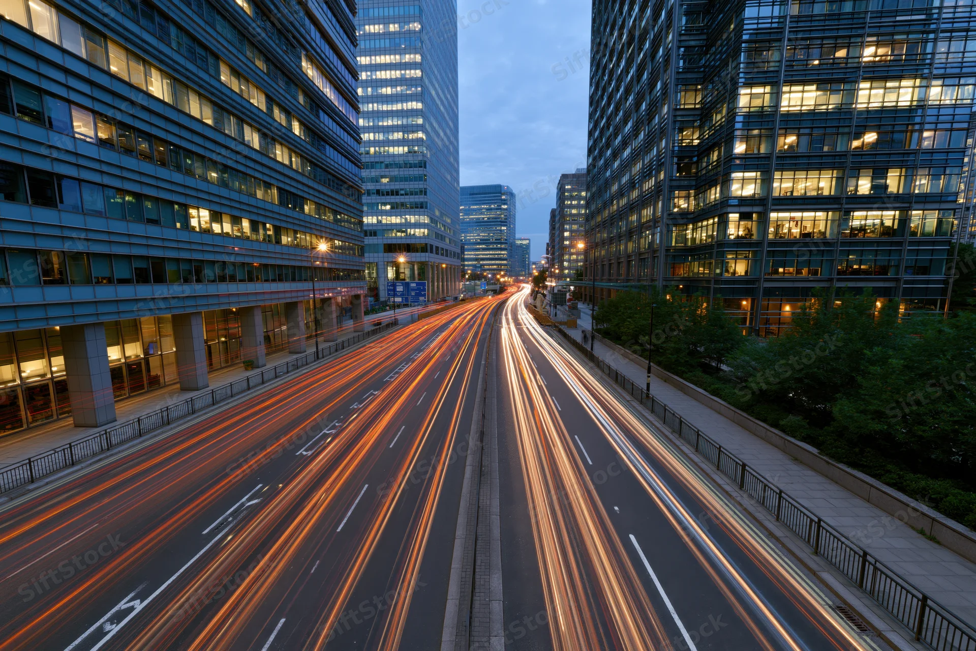 Long-exposure view of vehicle light trails moving along a multi-lane highway between modern glass skyscrapers in a city at dusk