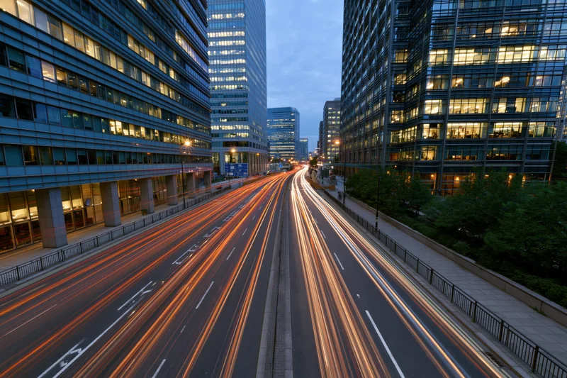 Long-exposure view of vehicle light trails moving along a multi-lane highway between modern glass skyscrapers in a city at dusk