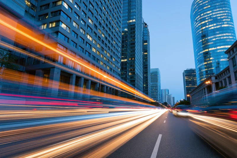 Vehicle light trails moving along a city street at dusk between tall glass skyscrapers