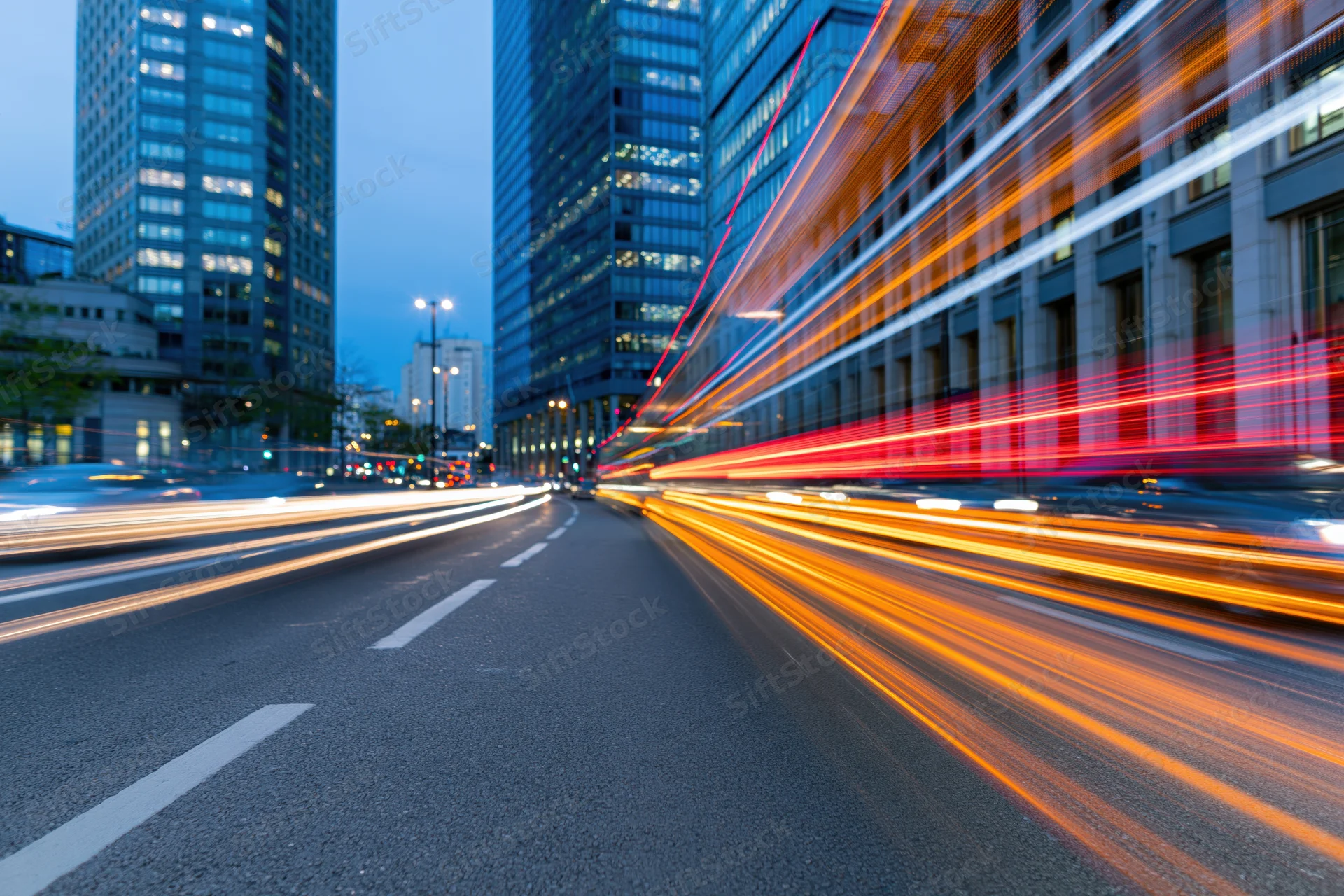 Light trails from moving vehicles on a city street at dusk between tall glass buildings