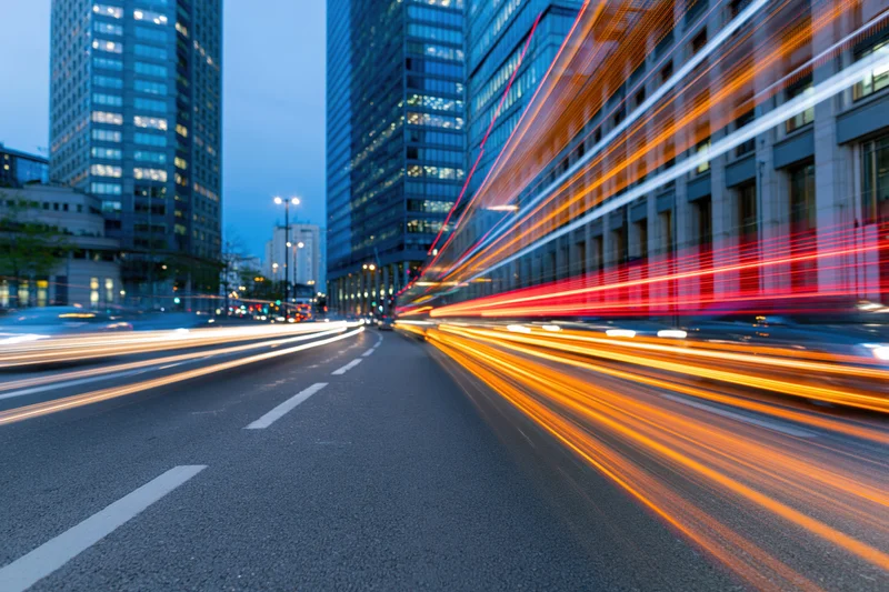 Light trails from moving vehicles on a city street at dusk between tall glass buildings