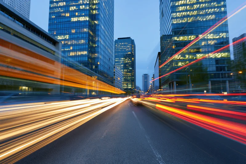 Light trails from moving vehicles on a city street at twilight between tall glass office buildings