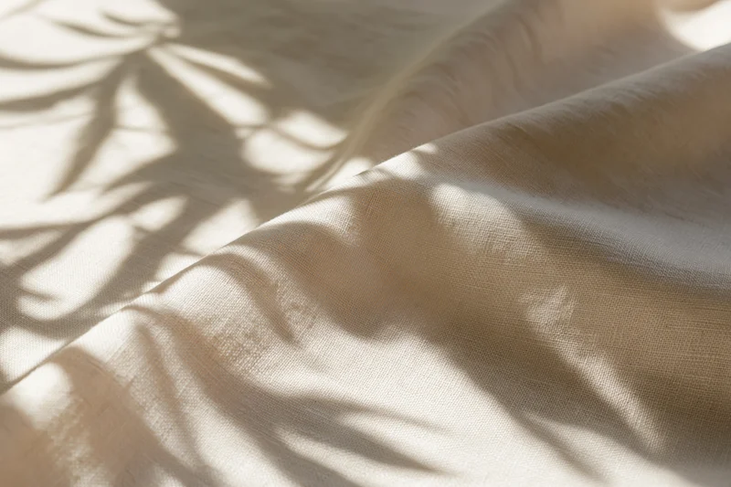 Natural linen fabric with soft diagonal folds and leaf shadows on a light surface