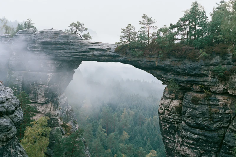 Natural sandstone rock arch spanning a misty gorge with sparse pine trees on the cliffs