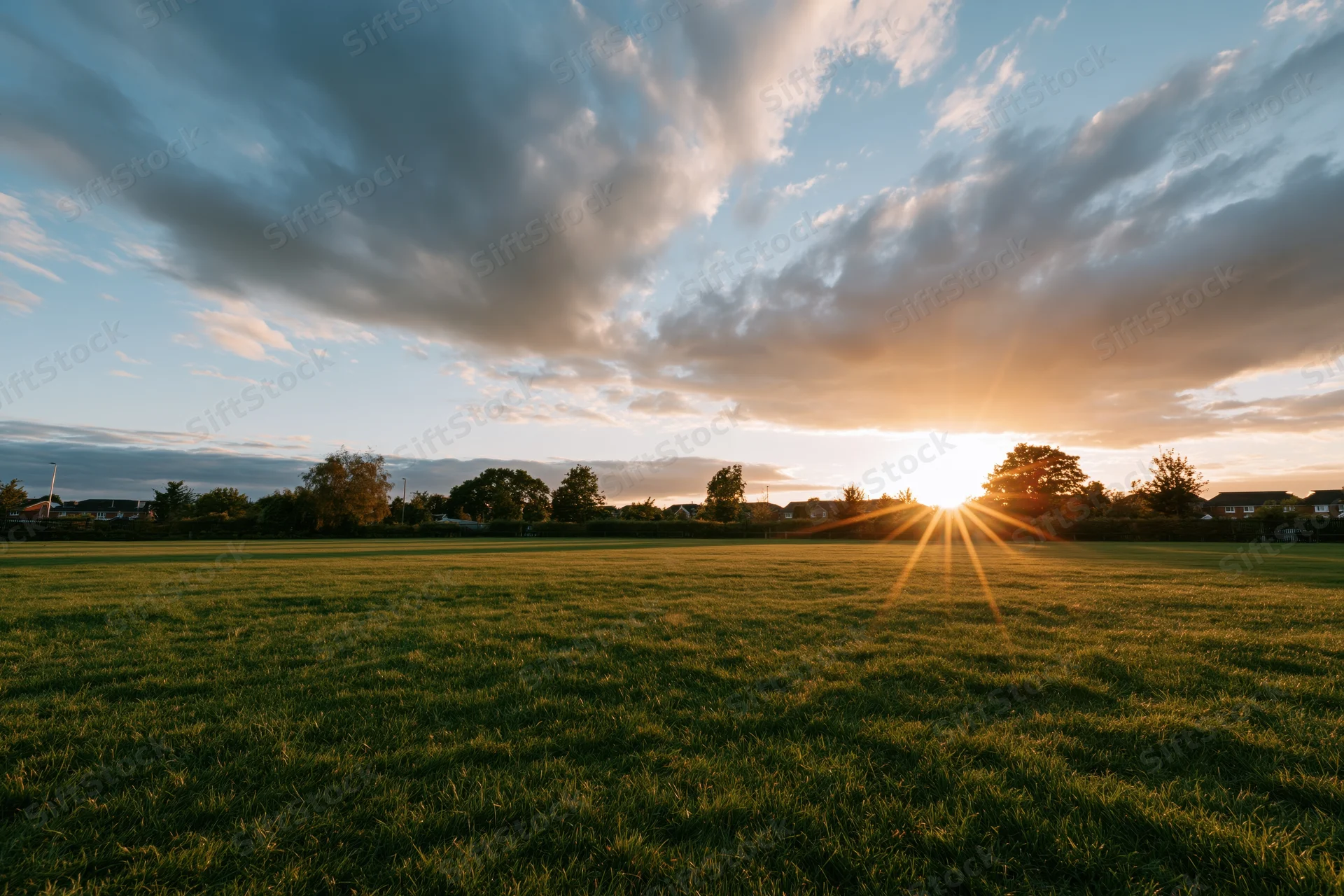 Open green meadow at golden hour with sun low on horizon casting rays through clouds