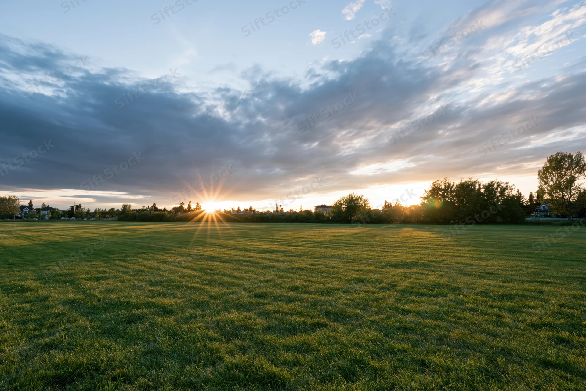 Open green meadow at golden hour with sun low on the horizon breaking through clouds