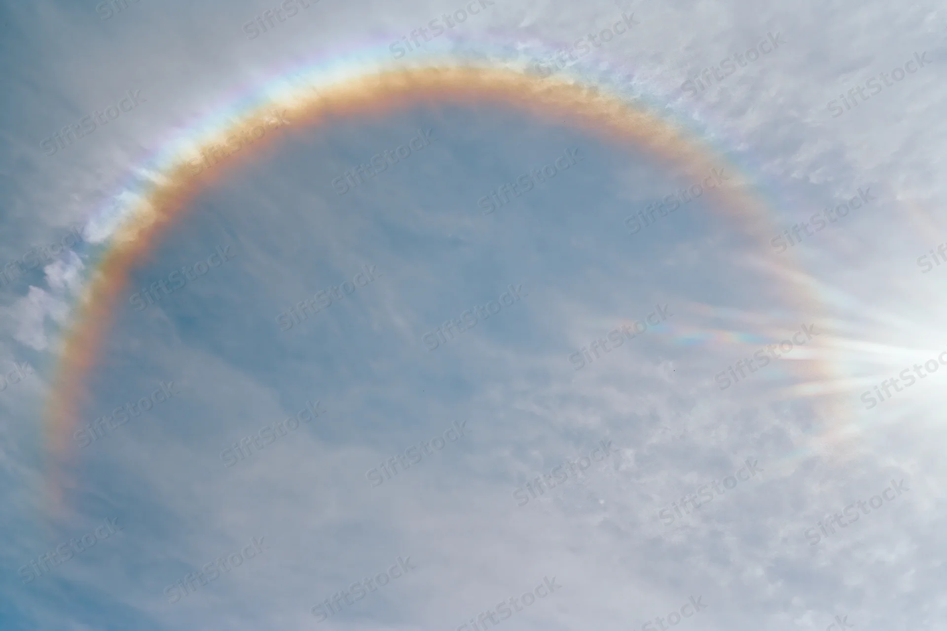 Overexposed daytime sky with wispy clouds and a rainbow arching upward, viewed from below