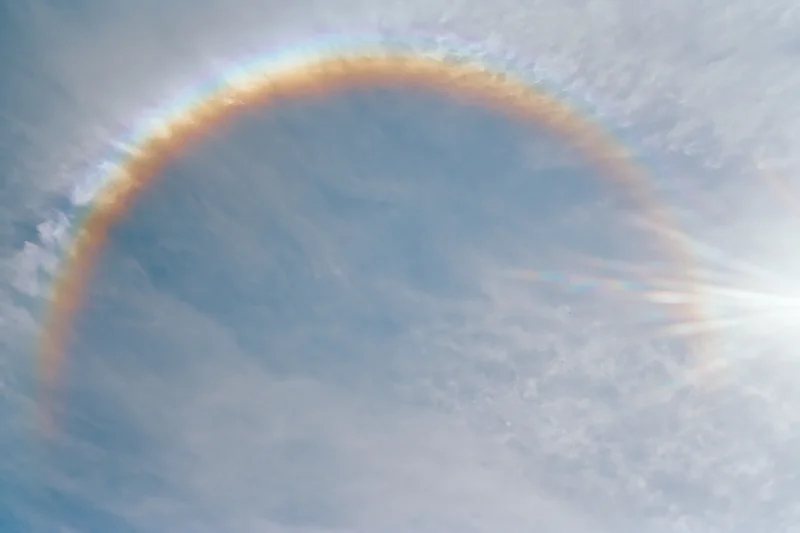 Overexposed daytime sky with wispy clouds and a rainbow arching upward, viewed from below
