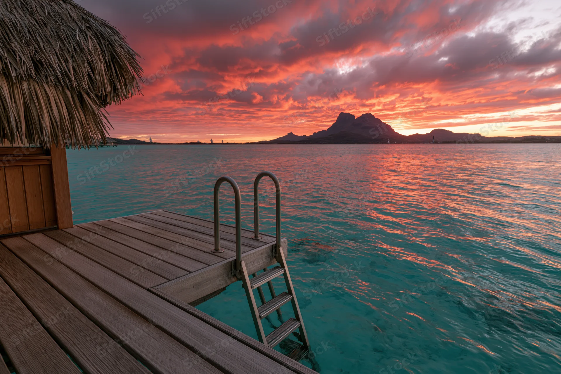 Overwater bungalow with thatched roof and wooden deck overlooking turquoise ocean at sunset