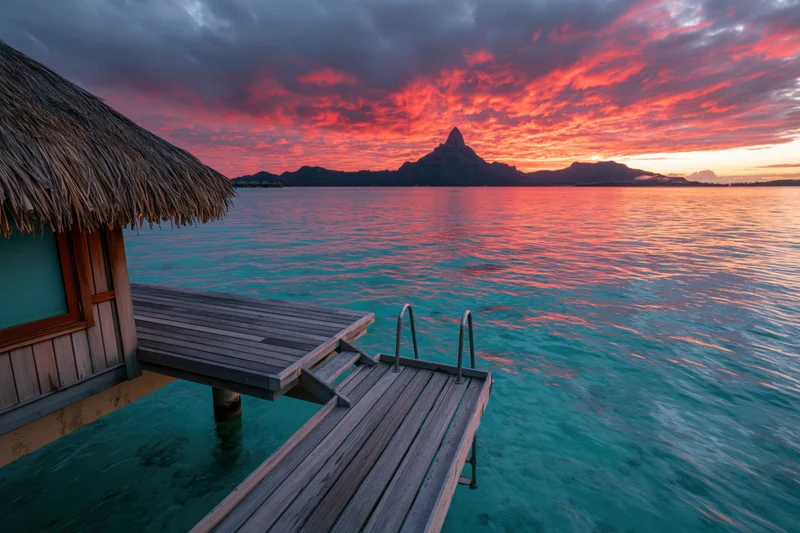 Overwater bungalow with thatched roof and wooden deck extending into turquoise ocean at sunset with distant mountain silhouette