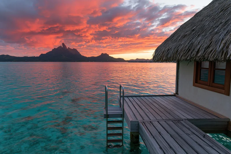 Overwater bungalow with thatched roof and wooden deck extending over turquoise water at sunset with distant mountains under pink and orange sky