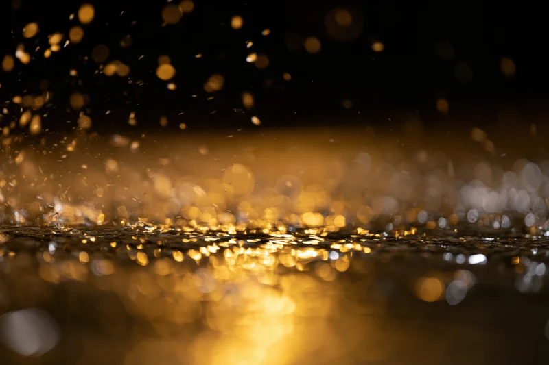 Raindrops falling on a dark wet surface at night creating golden reflections and bokeh lights