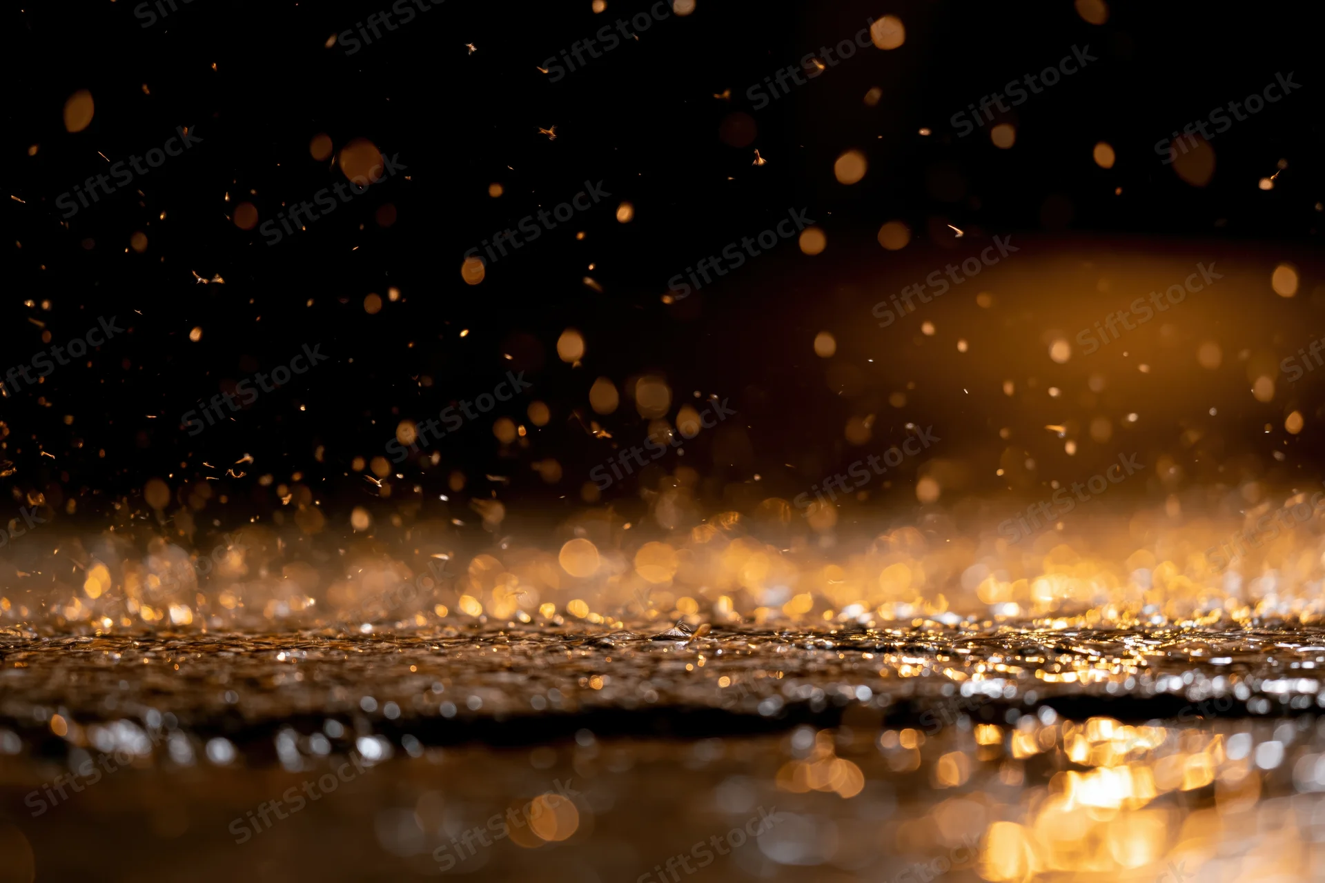 Rain falling on a wet dark surface at night creating golden reflections and bokeh lights