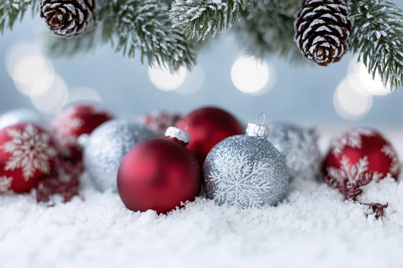 Red and silver christmas ornaments with snowflake patterns resting in snow beneath snowy pine branches