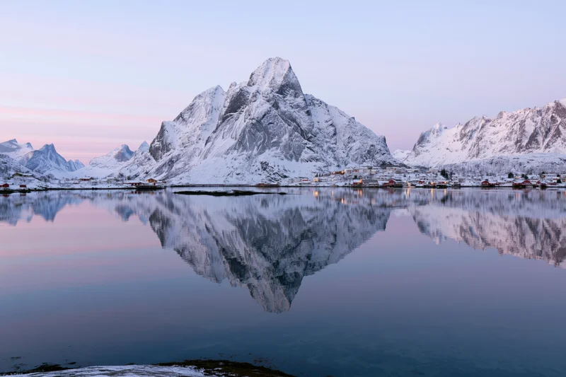 Snow-covered mountains reflected in calm water at twilight in reine fishing village, lofoten islands, norway