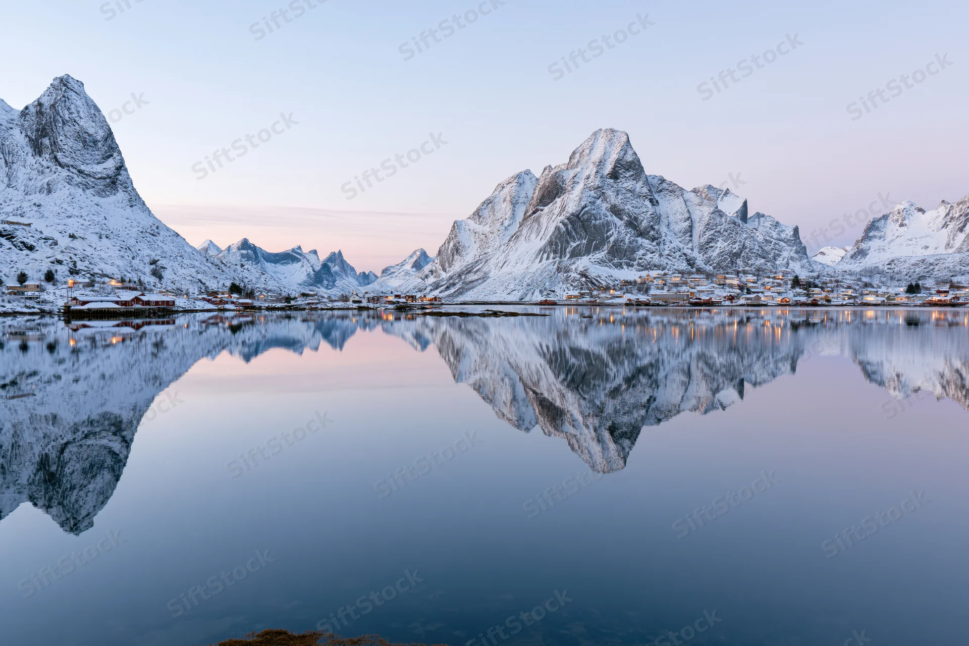 Snow-covered mountains and fishing village reflected in calm water at twilight in Reine, Lofoten, Norway