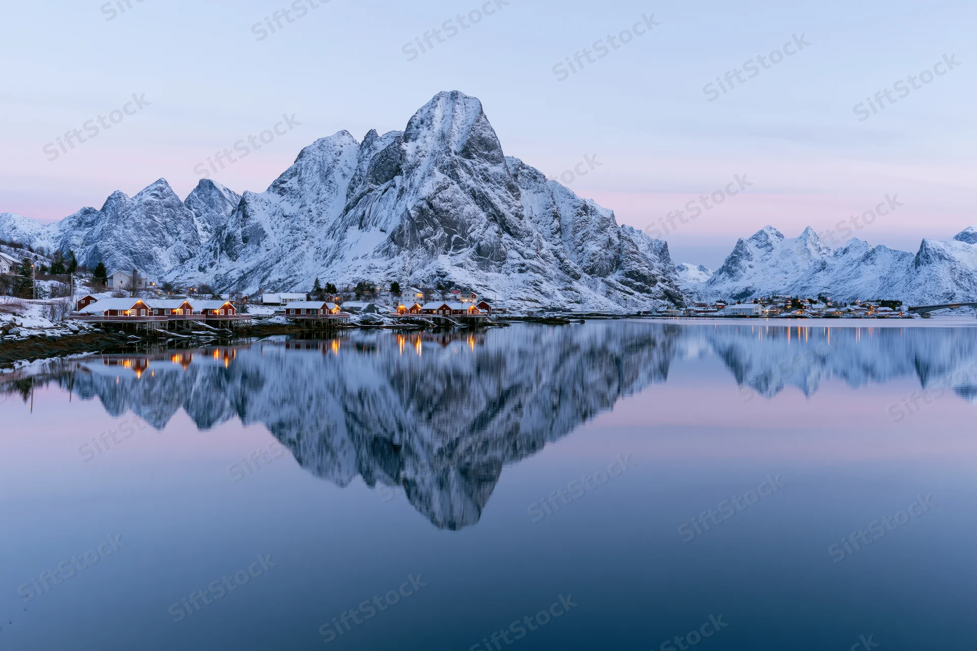 Fishing village cabins along a calm fjord reflecting snow-covered mountains at winter twilight in reine lofoten norway