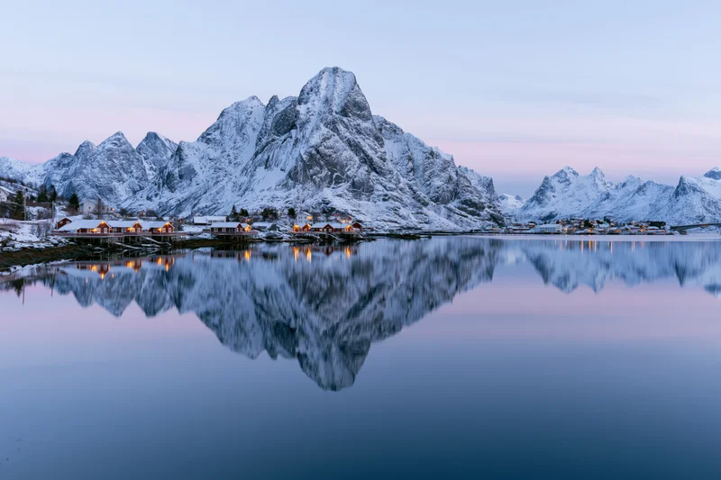 Fishing village cabins along a calm fjord reflecting snow-covered mountains at winter twilight in reine lofoten norway