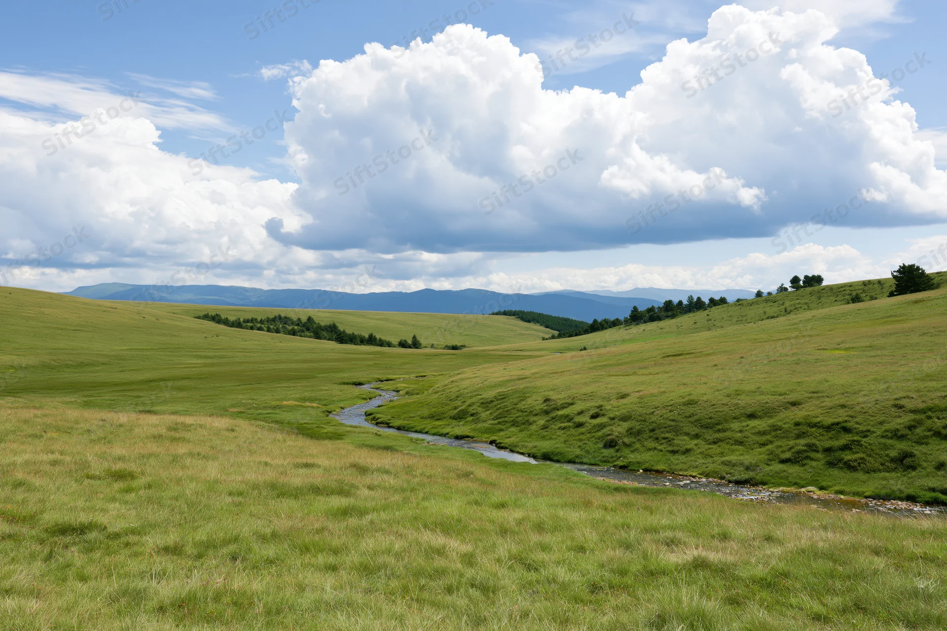 Rolling green meadow hills with a winding stream under a cloudy blue sky