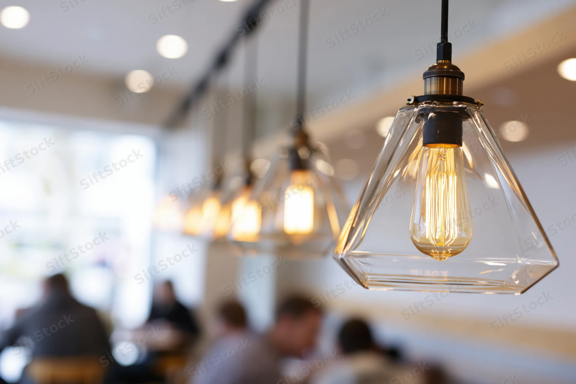 Row of geometric pyramid-shaped clear glass pendant lights hanging in a cafe interior with blurred diners below