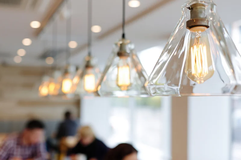 Row of geometric pyramid-shaped clear glass pendant lights hanging in a restaurant interior with warm glow and blurred diners below