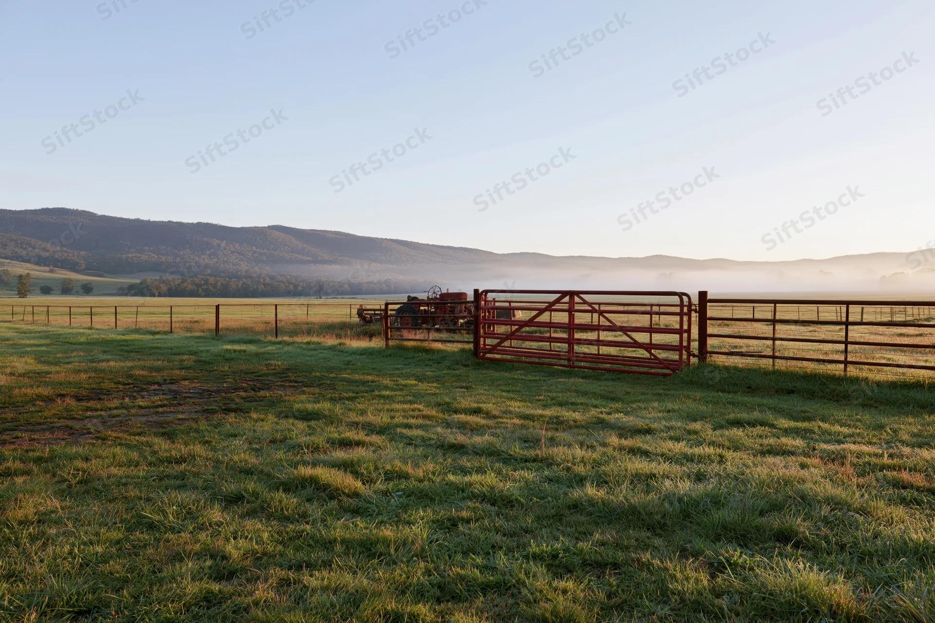 Open green pasture bordered by red metal gate and fence with tractor in background at sunrise