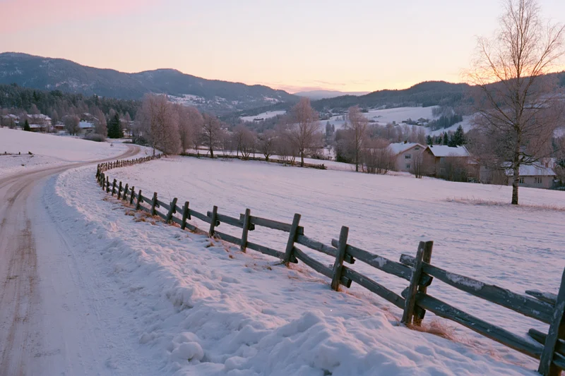Snow-covered rural road winding past a weathered wooden splitrail fence in a mountain valley at sunset