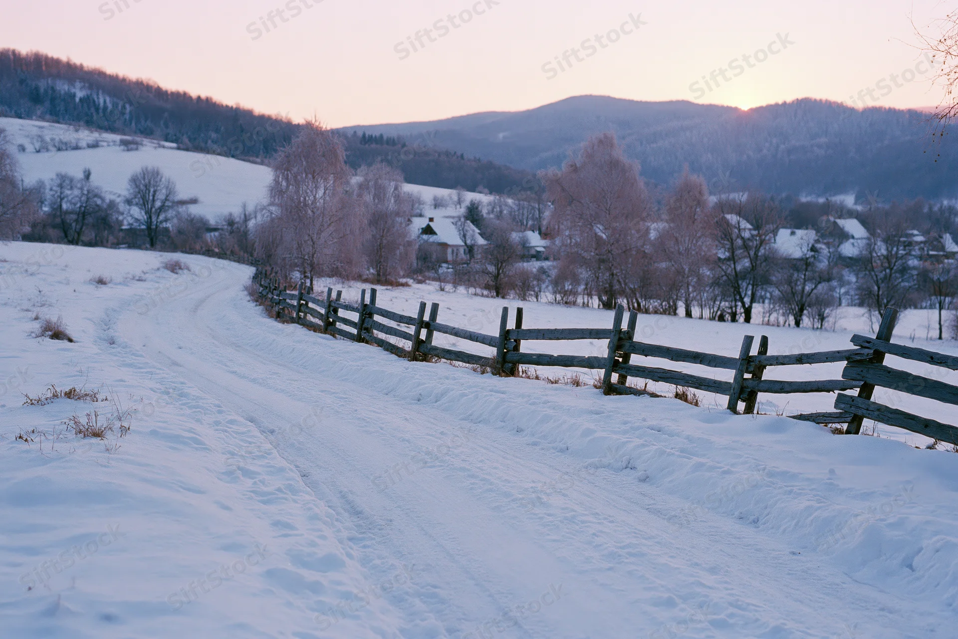 Snow-covered rural road winding past a weathered wooden split-rail fence in a mountainous countryside at twilight