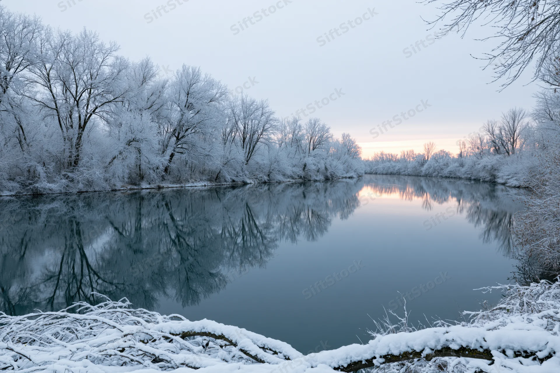 Snow-covered trees lining a still river in deep winter with reflections at dawn