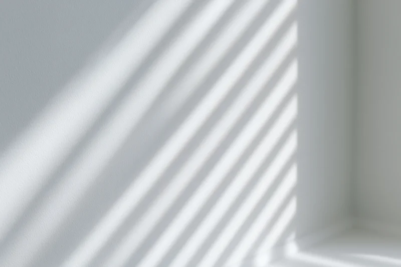 Diagonal shadows from window blinds cast across a white interior wall corner