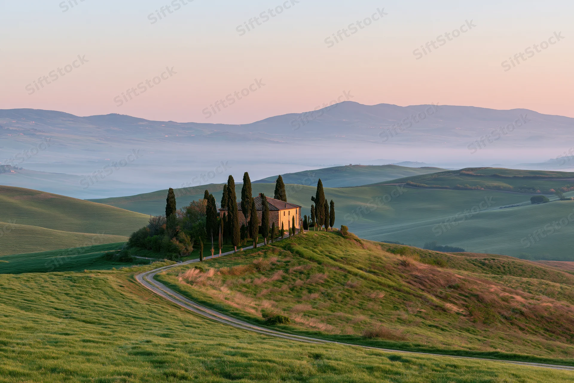 Stone farmhouse surrounded by tall cypress trees on a rolling green hill at sunset