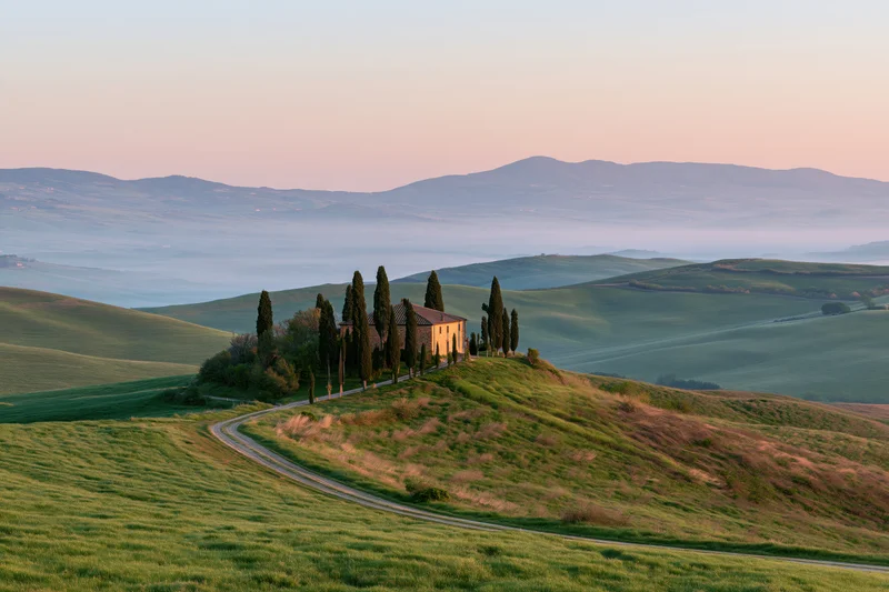 Stone farmhouse surrounded by tall cypress trees on a rolling green hill at sunset