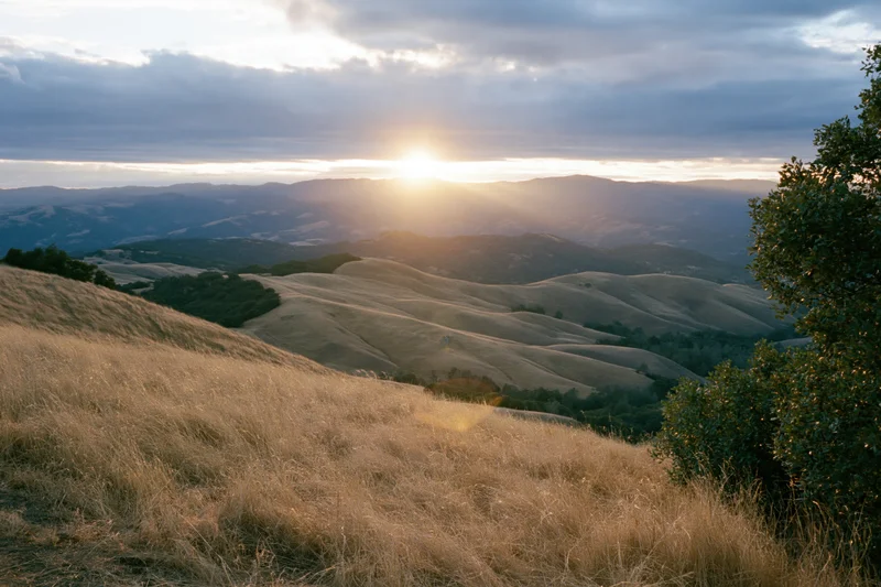 Sun setting behind rolling hills in california landscape with golden grass and distant mountains