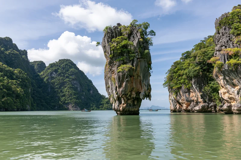 Tall limestone karst pillar rising from calm tropical bay surrounded by forested cliffs and small boats