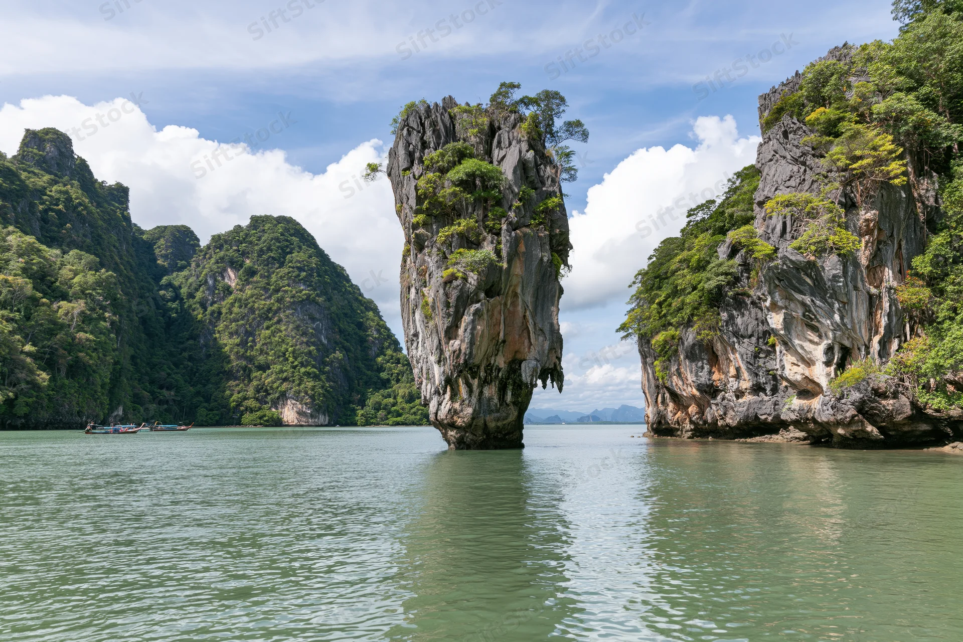 Tall limestone karst pillar rising from shallow tropical bay surrounded by forested cliffs and calm water