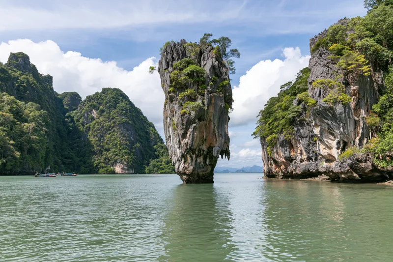 Tall limestone karst pillar rising from shallow tropical bay surrounded by forested cliffs and calm water