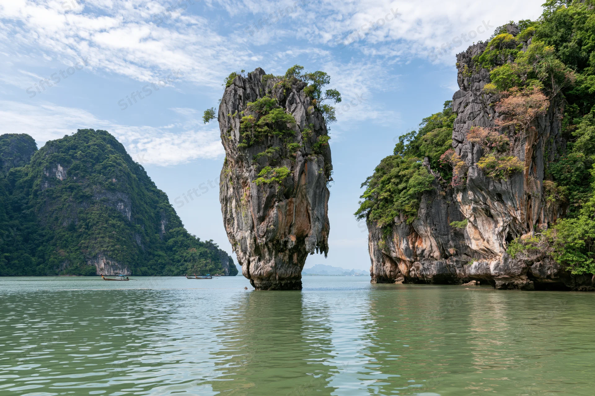Tall limestone karst pillar rising from shallow tropical bay surrounded by forested cliffs and calm water with boats in the distance