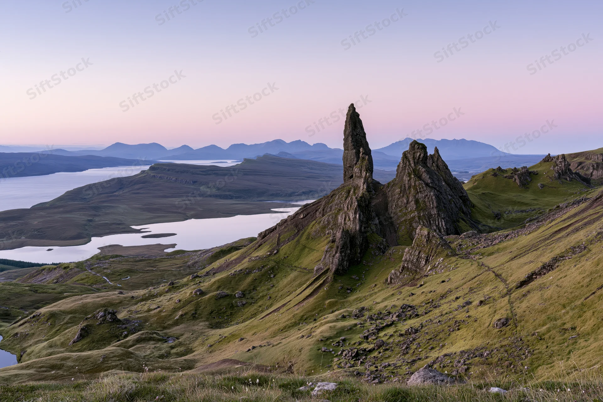 Old Man of Storr rock pinnacle rising from green hillside overlooking loch and distant mountains at twilight